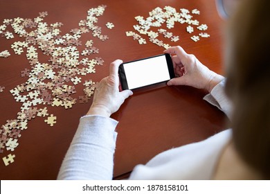 Old Woman Looking At Smartphone While Doing A Jigsaw Puzzle