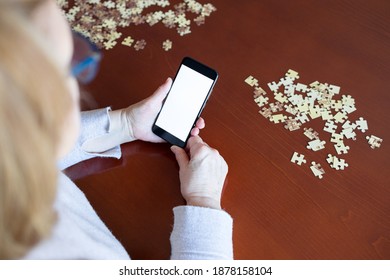 Old Woman Looking At Smartphone While Doing A Jigsaw Puzzle