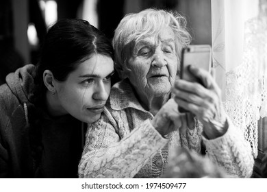 An Old Woman Looks At A Smartphone, With His Adult Granddaughter. Black And White Photo.