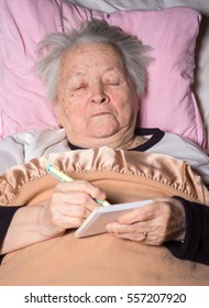 Old Woman Lying In Bed And Making Notes At Home