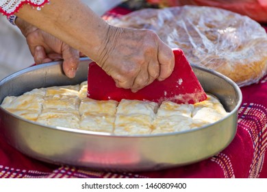 Old woman making traditional bulgarian pie with white cheese called banitsa. Cutting the pieces with old tool.