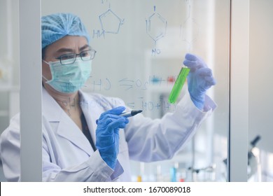 Old woman medical research scientist holding the green chemical tube and write down the chemical formula on the glass board