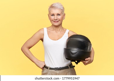 Old Woman With Motorbike Helmet In Studio On Yellow Background Have Smile On Her Face. Extreme Biker With Tender View. Minimal Stylish 50s Rock Lady.