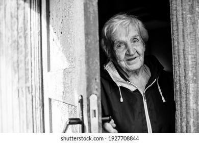 An Old Woman Peeks Out From Behind The Door Of His House. Black And White Photo.