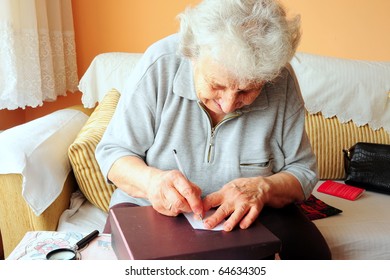 Old Woman, Portrait Of Old Senior Woman Writing Something On White Paper With A Pen On The Table In Living Room At Home