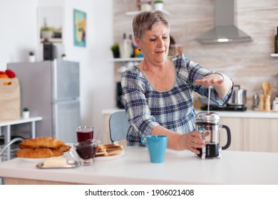 Old woman pushing down the lid of french press while making coffee for breakfast in kitchen. Elderly person in the morning enjoying fresh brown cafe espresso caffeine from mug filter relax refreshment - Powered by Shutterstock