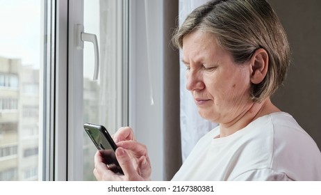 Old Woman Scrolls Through Web Pages On Black Smartphone Standing Near Window. Woman In White T-shirt Checks Social Media On Mobile Phone