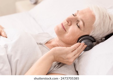 Old woman is seen reclining in a bed, wearing headphones. She appears relaxed, listening to music or a podcast as she rests comfortably. - Powered by Shutterstock