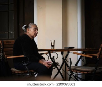 An Old Woman Sits Alone And Pensive At A Bar Table Outside. A Glass Of Drink Is On The Table. Woman Alone With Her Thoughts. France, Paris. September, 29, 2022