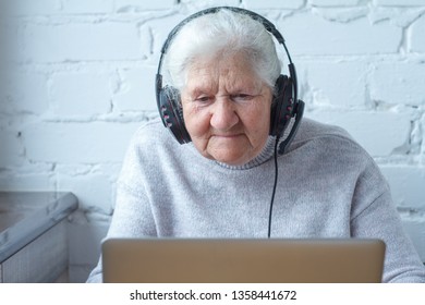 An old woman sits at a table in front of a laptop with headphones. Education for adults. Freelancer works. Retired woman is chatting, blogging, checking email. Social media, network.