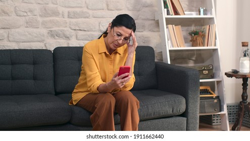 The old woman, sitting on the sofa at home, reading the news on her mobile phone, is upset by the news she sees.