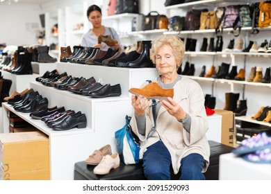 Old Woman Sitting In Salesroom Of Shoeshop And Choosing New Shoes. Asian Woman Shopping In Background.