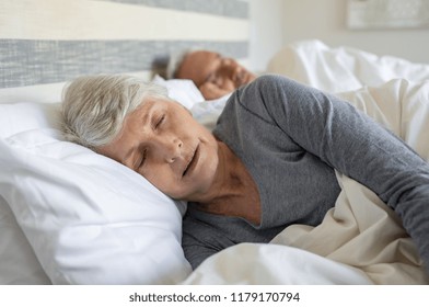 Old Woman Sleeping On Bed At Home With Her Husband. Elder Lady Sleeping In The Bedroom With Husband In Background. Senior Woman With Grey Hair Wearing Nightwear Asleep In Bed.