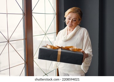 Old Woman Standing Indoors Holding A Gift Box