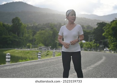 An old woman stands on a peaceful road with scenic mountain views, taking a moment to pause and reflect during her outdoor workout. - Powered by Shutterstock