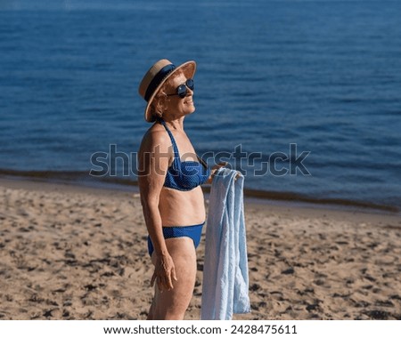 An old woman in a straw hat, sunglasses and a swimsuit is resting on the beach. 
