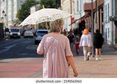 Old Woman With Sun Umbrella Walk On A Street On People Background. Hot Weather, Life Of Elderly People In Summer