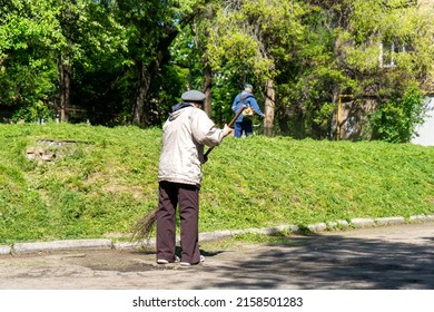 Old Woman Sweeping The Street In The City. Man In The Background Mows The Grass