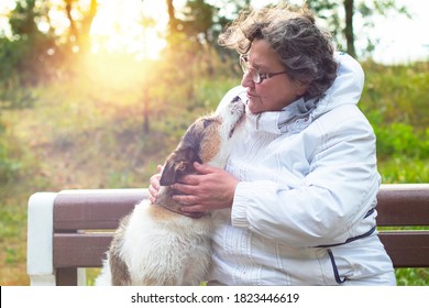 An Old Woman Is Talking To Her Pet. Portrait Of An Elderly Man Hugging Her Dog And Kissing An Animal In The Park On A Bench.