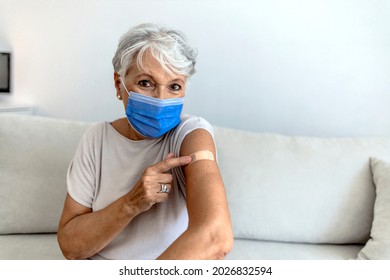 Old Woman That Has Received Her COVID-19 Vaccine. She Is Pulling Up Her Sleeve To Show The Bandage. Happy Female Wearing Protective Face Mask Looking At Camera After Receiving Coronavirus Vaccination.