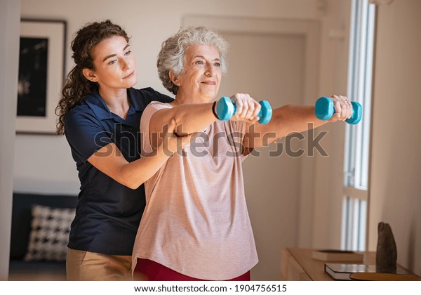 Old woman training with physiotherapist using\
dumbbells at home. Therapist assisting senior woman with exercises\
in nursing home. Elderly patient using dumbbells with outstretched\
arms.