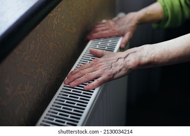 An Old Woman Tries To Warm Her Hands On A Cold Radiator Indoors