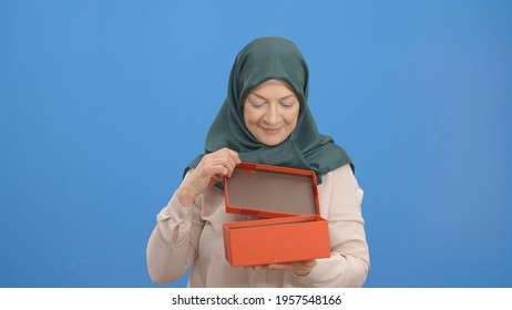 Old Woman In A Turban Opens Gift Box, Isolated On White Background Studio Woman. The Woman Is Surprised By The Gift In The Box.