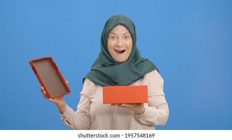 Old Woman In A Turban Opens Gift Box, Isolated On White Background Studio Woman. The Woman Is Surprised By The Gift In The Box.