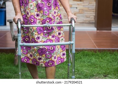 Old Woman Walk Outdoors By Walking Frame After Knee Joint Replacement Surgery With Suture Scar On Left Leg. Physical Therapy Recovery On Grass Field