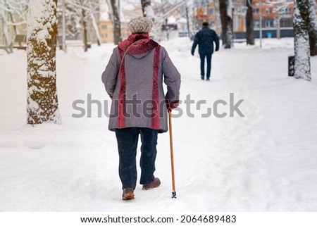 Old woman walking along slippery winter road with walking stick. Grandma with cane walking along snowy street. Senior woman with walking stick in hands walks along snowy path.