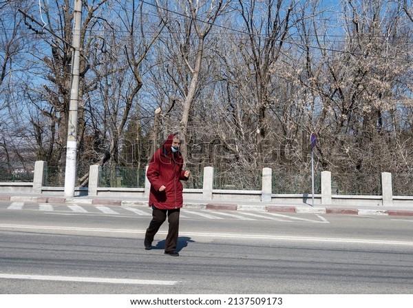 An old woman walks across the\
street in a hurry. The woman crosses the street in insecurity - she\
does not walk on the crosswalk. Romania, Gorj. March, 18,\
2022