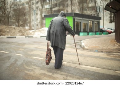 Old Woman Walks Down Road In Black Jacket. Pensioner In Russia On Street. Grandmother With Walking Stick.