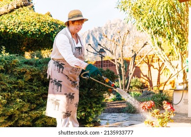 Old Woman Watering The Plants In Her Garden With A Hosepipe