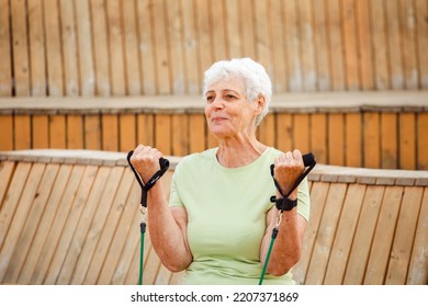 Old woman wear green t-shirt doing exercises outdoor , sitting on wooden bench using resistance rubber bands. Healthy lifestyle, active retired life and sporty time. - Powered by Shutterstock