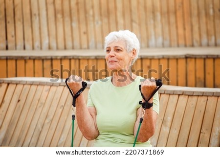 Old woman wear green t-shirt doing exercises outdoor , sitting on wooden bench using resistance rubber bands. Healthy lifestyle, active retired life and sporty time.