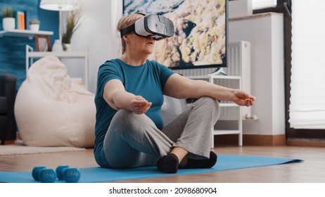 Old woman wearing vr glasses and doing meditation while sitting in lotus position on yoga mat. Senior person using virtual reality goggles to meditate for wellness and recreation. - Shutterstock ID 2098680904