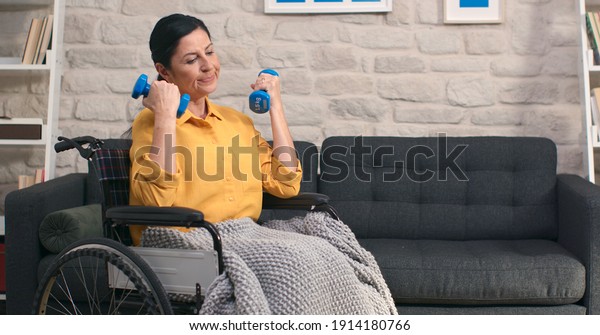 Old woman in wheelchair\
exercising and lifting weights in a living room. Rehabilitation of\
young woman in wheelchair. Home exercise, fitness, rest and\
lifting.