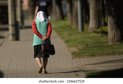 An Old Woman In A White Headscarf, A Turquoise Jacket With A Bag In Her Hands Walks Down The Street