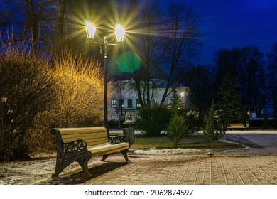 Old wooden bench dusted with first snow is illuminated by street lamp. City park in evening