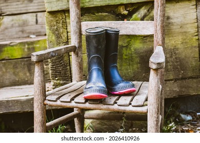 A Old Wooden Chair With Some Kids Black Rubber Boots Sitting In It- Black Water Boots Sitting In A Antique Brown Chair- Black Muck Boots On A Vintage Childs Chair