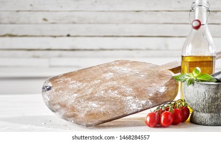 Old Wooden Empty Pizzeria Pizza Paddle With Flour On A White Wood Table Alongside Tomatoes, Olive Oil And A Pestle And Mortar With Copy Space