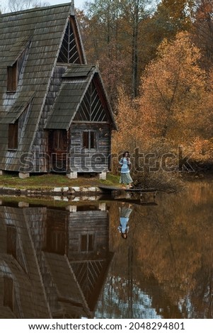 Old wooden house hut by lake among autumn forest, woman walking by wooden pier. Foliage reflection in water.