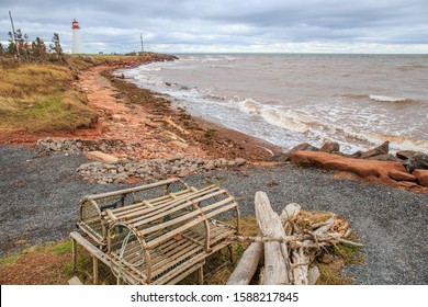 Old wooden lobster traps sit near a red sand beach and the white rolling waves of the Atlantic Ocean. A tall white and red lighthouse stands in the distance. 