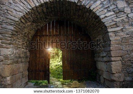 Old wooden open door in gates to ancient castle. Archway in stone fortress wall. Dark medieval woods doorway to summer sun light secret garden
