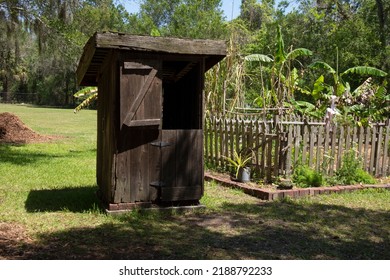 Old wooden outhouse by garden surrounded by wood panel fence with forest in the background on a beautiful sunny morning.