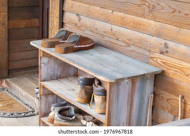 Old Wooden Shoebox Near A Wooden Cottage On A Farm. The Shoe Box Has Old Clogs, Rubber Boots And Shoes.