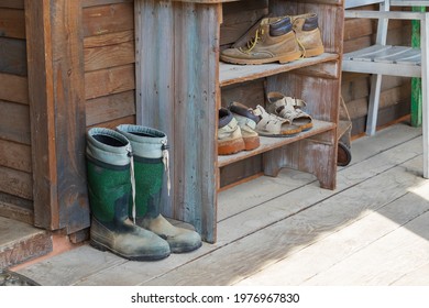 Old Wooden Shoebox Near A Wooden Cottage On A Farm. The Shoe Box Has Old Clogs, Rubber Boots And Shoes.