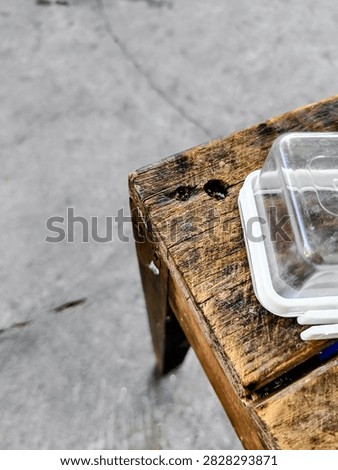 Old Wooden Stool with Clear Plastic Box on Concrete Floor  
Close up of the corner of an old weathered wooden stool with a small clear plastic box on top. Concrete floor background outdoor.
