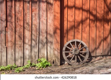 Old wooden wagon wheel reclining on red wooden wall. Wheel with boardwalk wall on background and cobbles on foreground. Place for your text. Wheel leaning up against wall. Sunny day. Skansen, Sweden