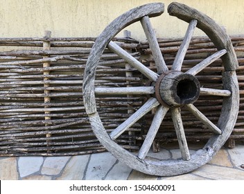 Old wooden wheel from a cart horse. The wheel of a wooden stroller since ancient times. Scenery in the yard: a wheel made of wood against the background of a fence made of branches.